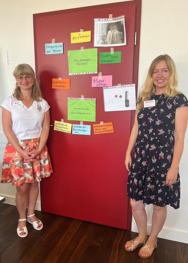 Bild vergrößern: Frau Beatrice Diewert und Frau Denise Lanitz stehen vor einer roten Türe, an der eine Art Mindmap mit Themen des Hospizdienstes abgebildet ist